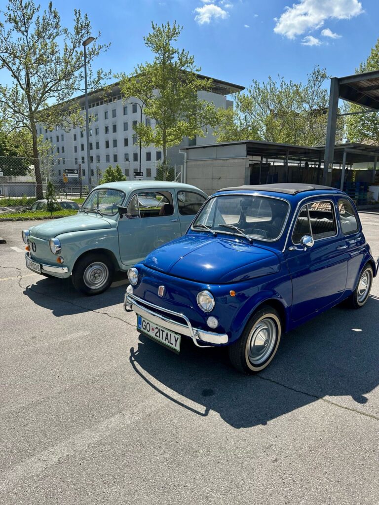 Luigi Aperitivo – klasični modri Fiat 500 parkiran ob sodobnih avtomobilih, simbol pristne osebnosti in italijanskega šarma.